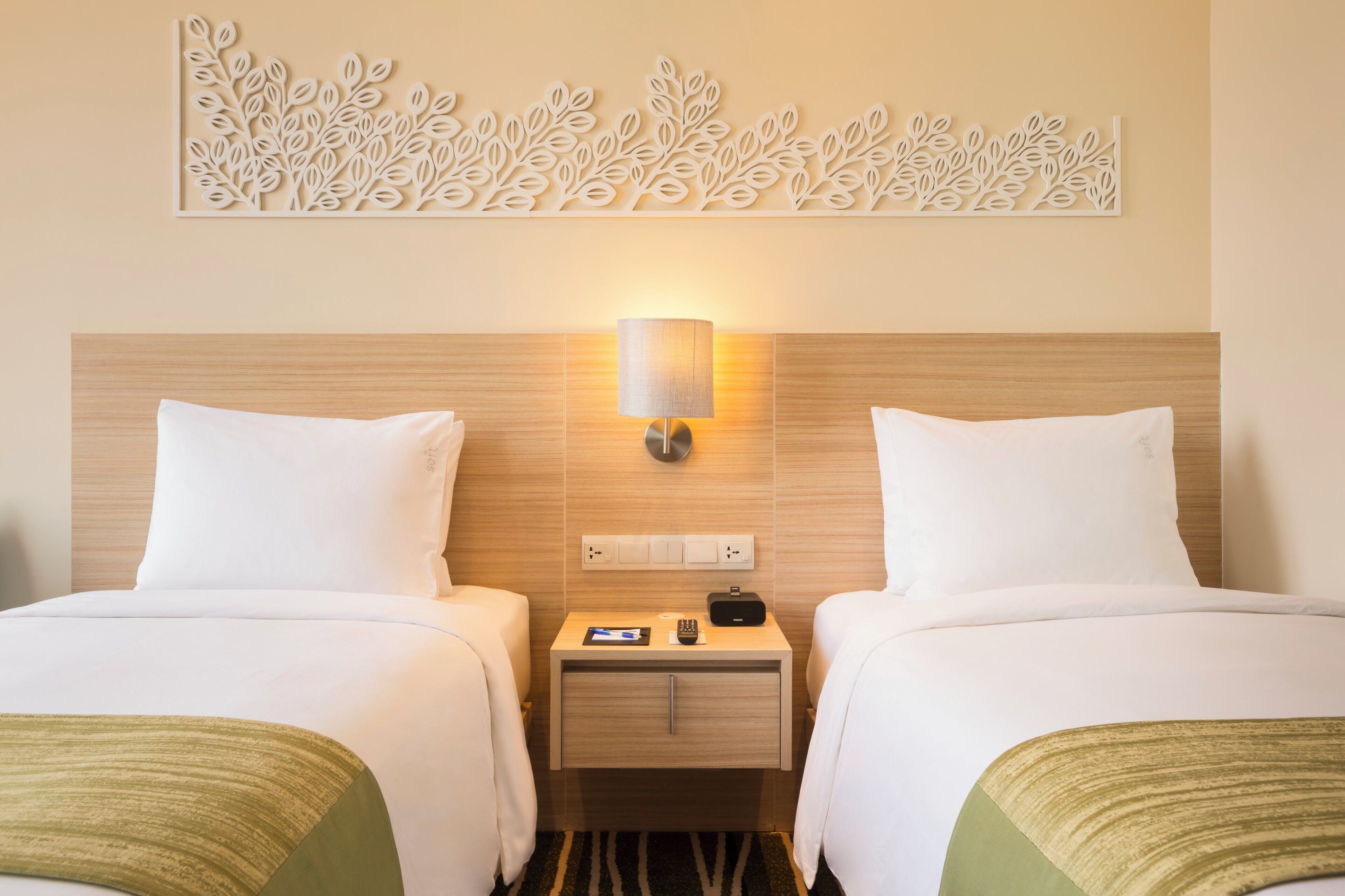Habitación de hotel con dos camas, una mesita de noche con lámpara, un adorno en la pared y un enchufe.
