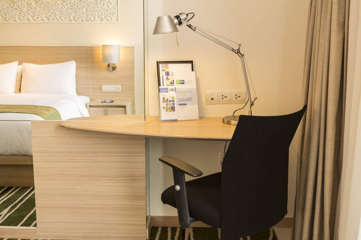 A hotel room with a bed, a desk with a lamp, a chair, and a wall-mounted power outlet.