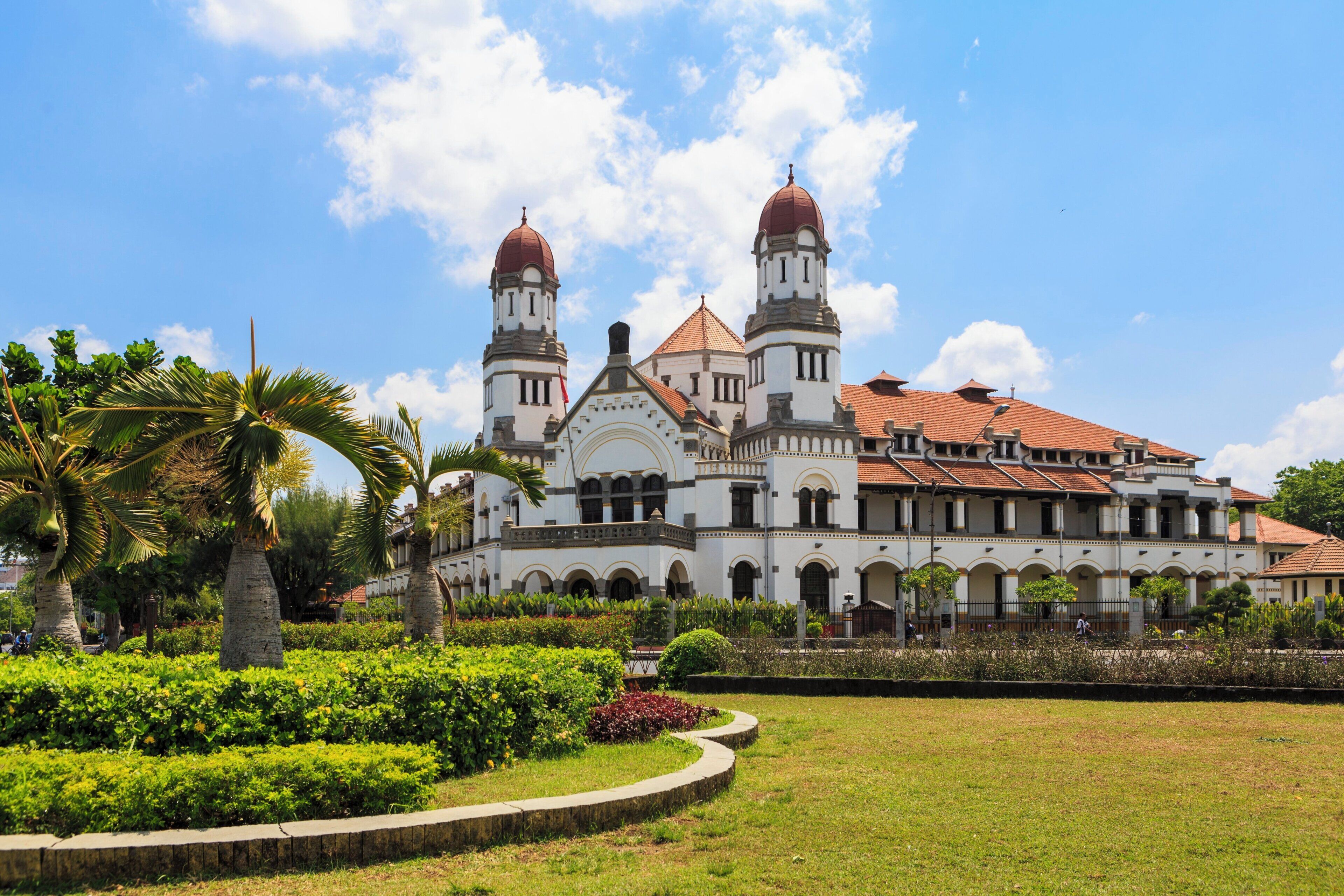 Un edificio grande e histórico con dos torres destacadas y una cúpula central, rodeado de jardines cuidados y palmeras.