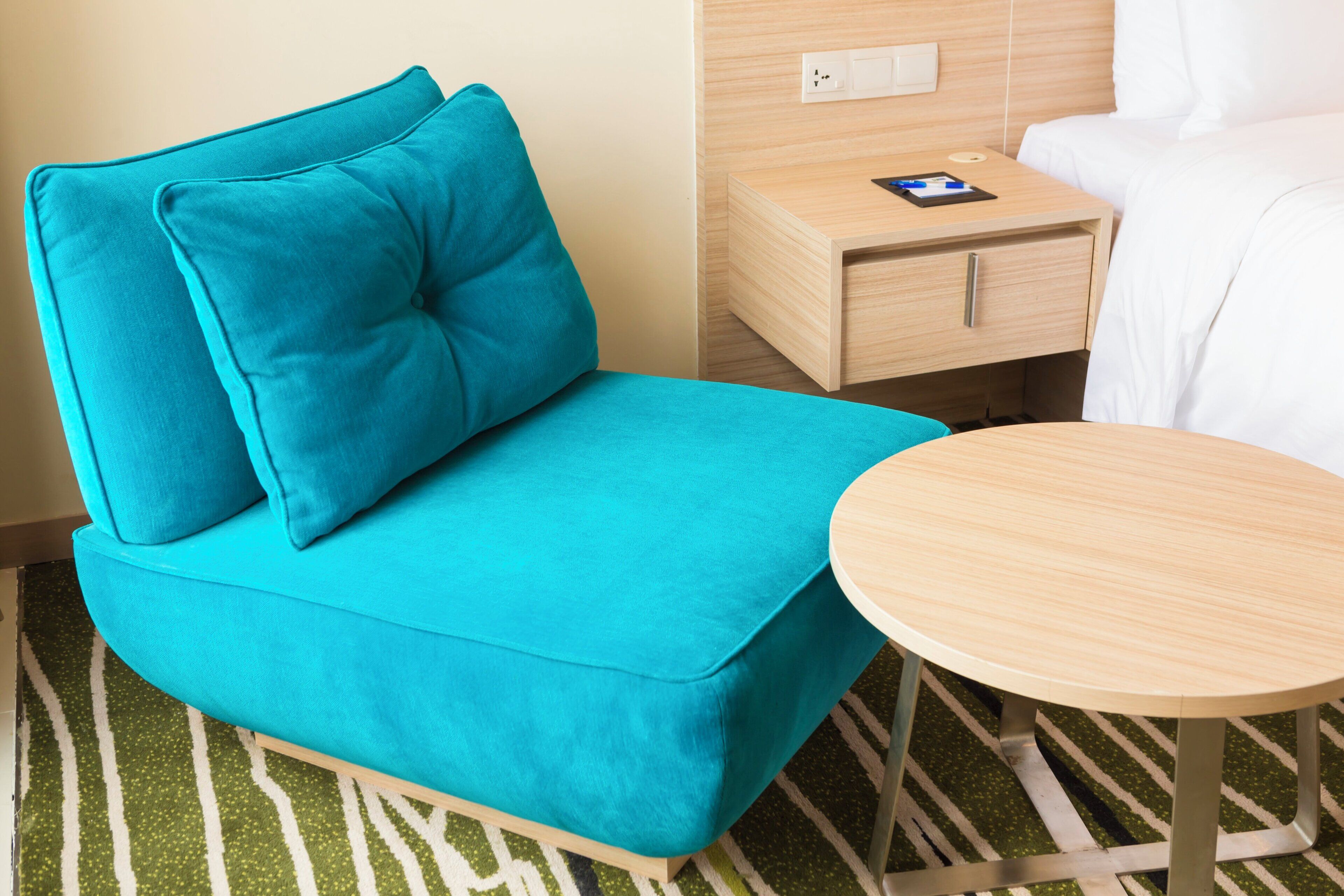 A teal sofa with a matching pillow, a wooden bedside table with a book and pen, and a round wooden table.