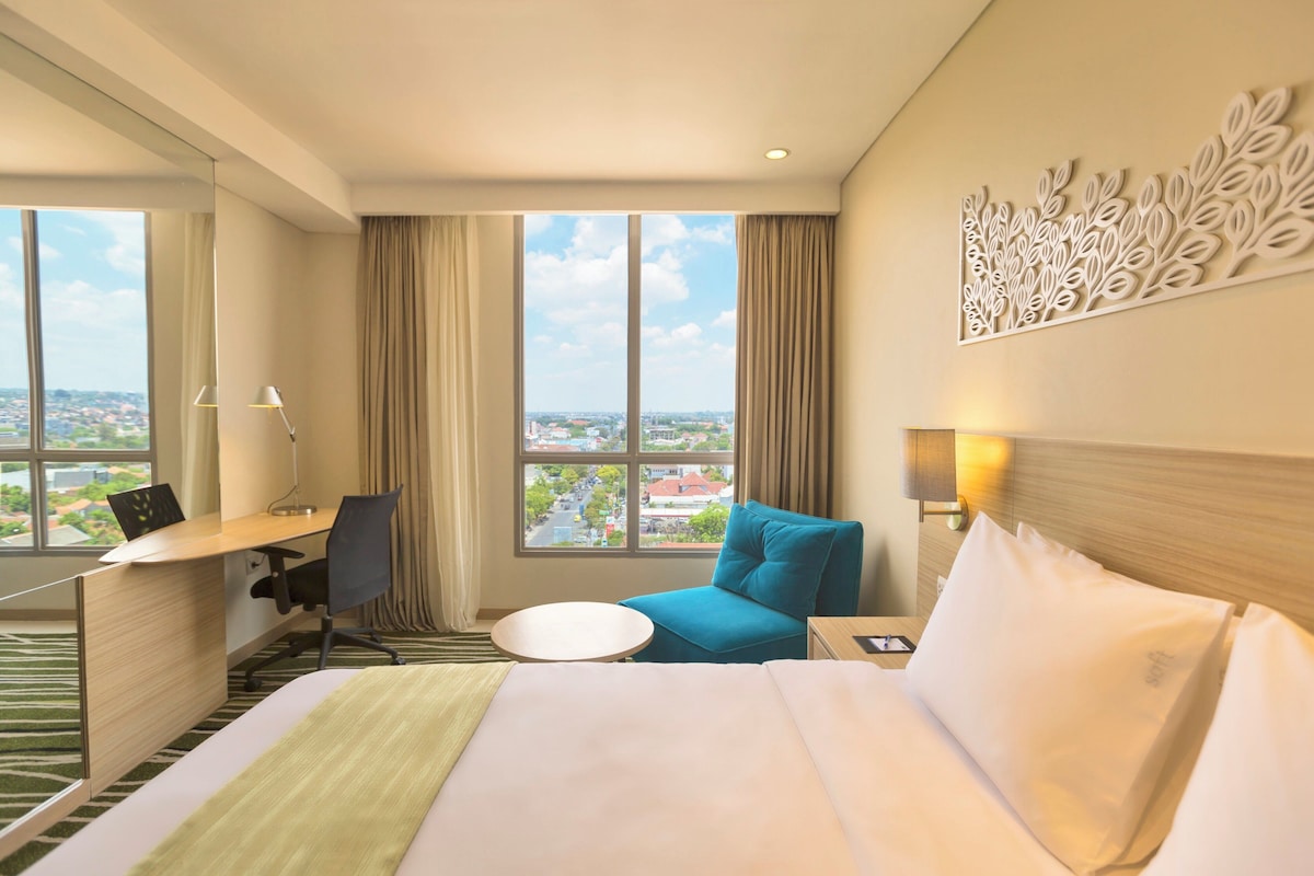A hotel room with a bed, desk, chair, and a view of the city.
