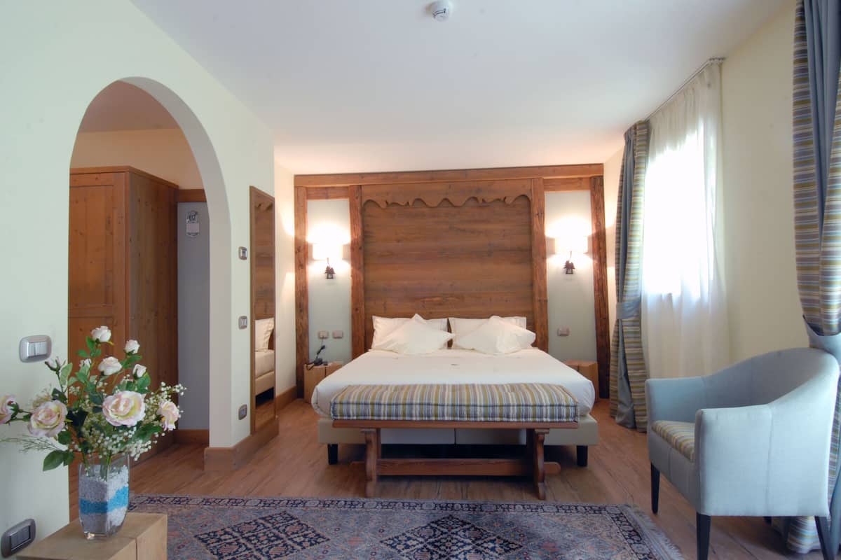 Une chambre à coucher avec une tête de lit en bois, un lit recouvert de draps blancs, un banc à rayures bleues et blanches, un fauteuil bleu clair et un vase rempli de fleurs posé sur une table d’appoint.