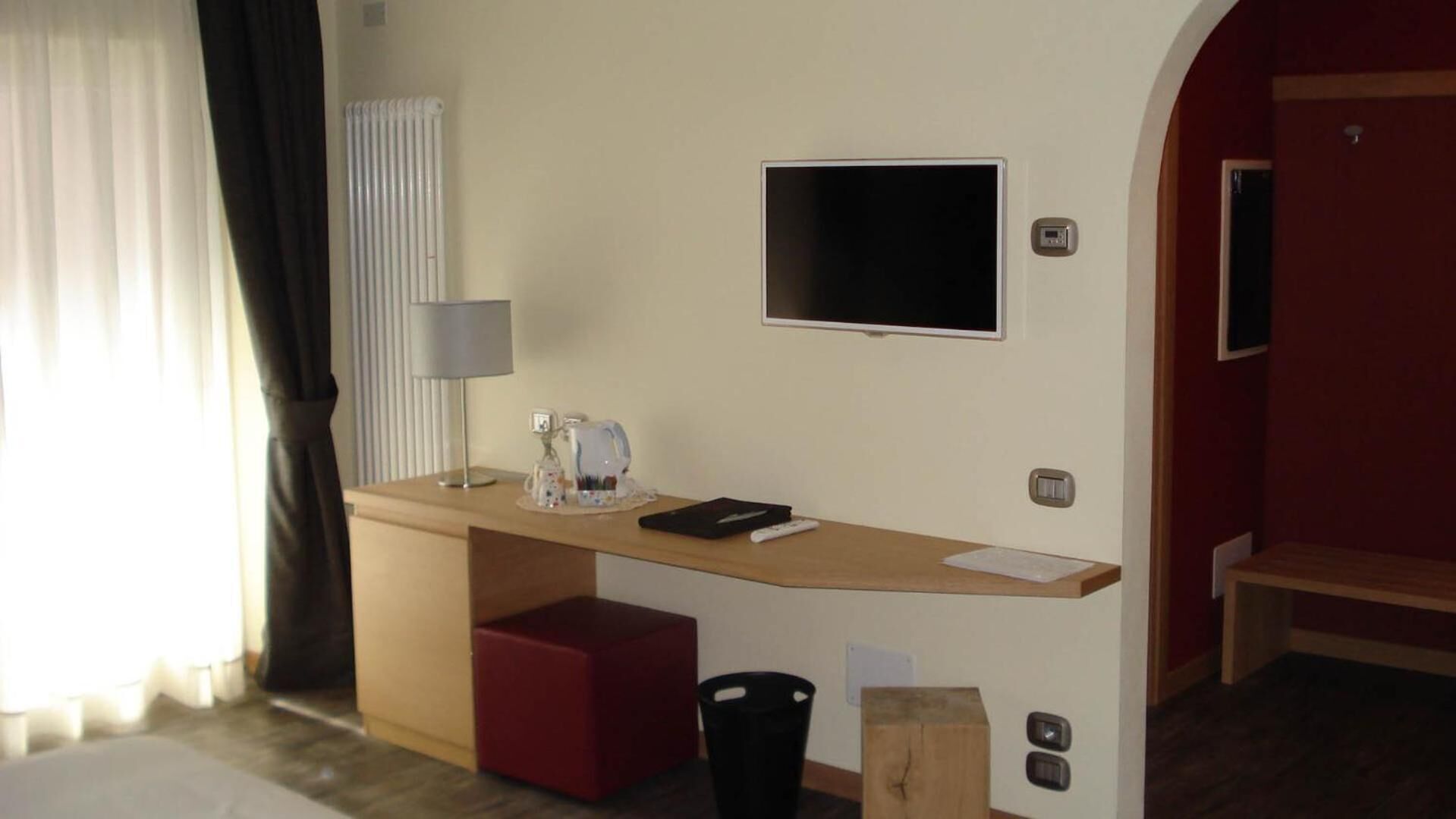 Quarto de hotel com uma escrivaninha de madeira, televisão de tela plana, luminária, um banquinho vermelho, lixeira e um pequeno banquinho de madeira.