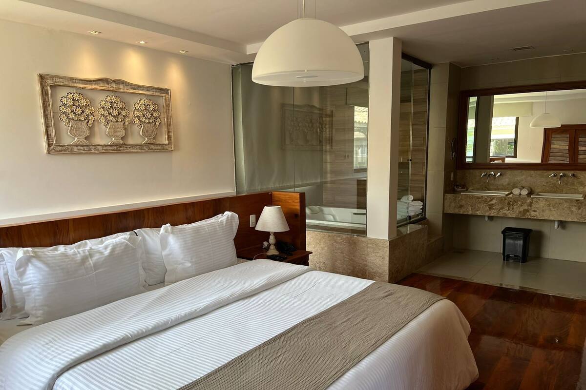 A hotel room with a large bed, a bedside table with a lamp, a glass shower enclosure, and a bathroom with a sink and mirror.