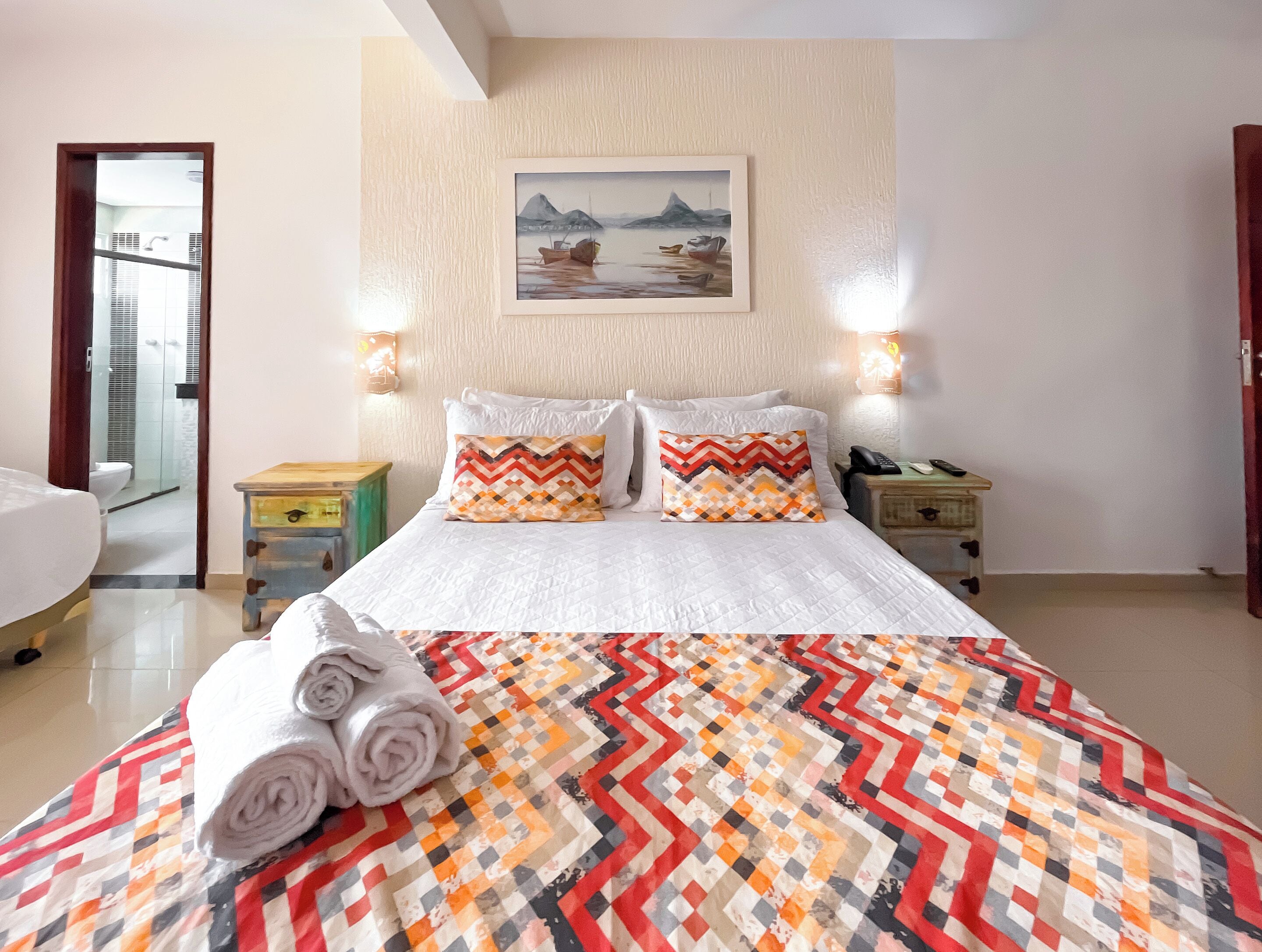 Une chambre d’hôtel avec un lit, deux tables de chevet, un tableau au mur et une salle de bain visible à travers une porte ouverte.