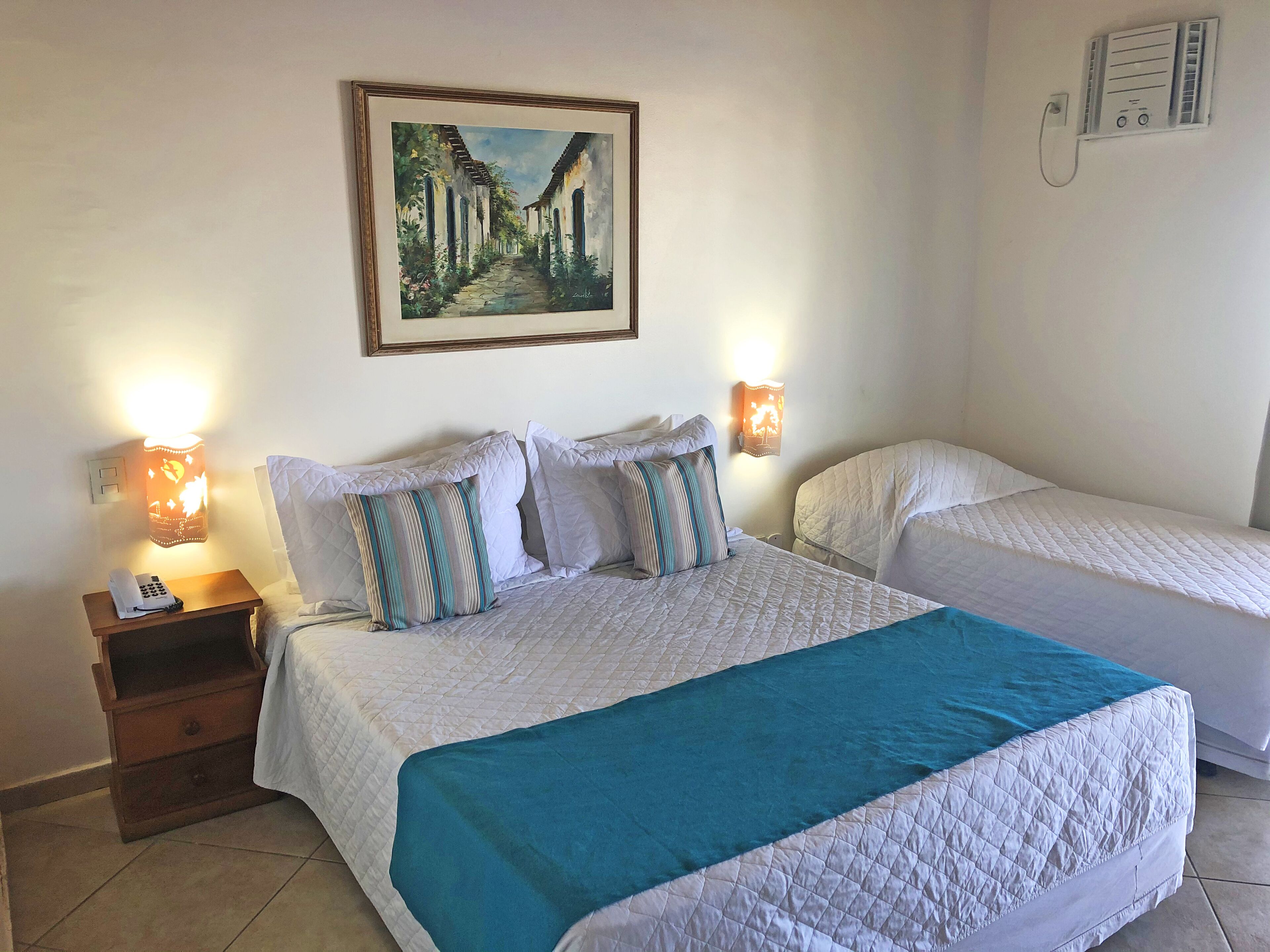 A hotel room with two beds, a nightstand with a phone, a painting on the wall, and a wall-mounted heater.