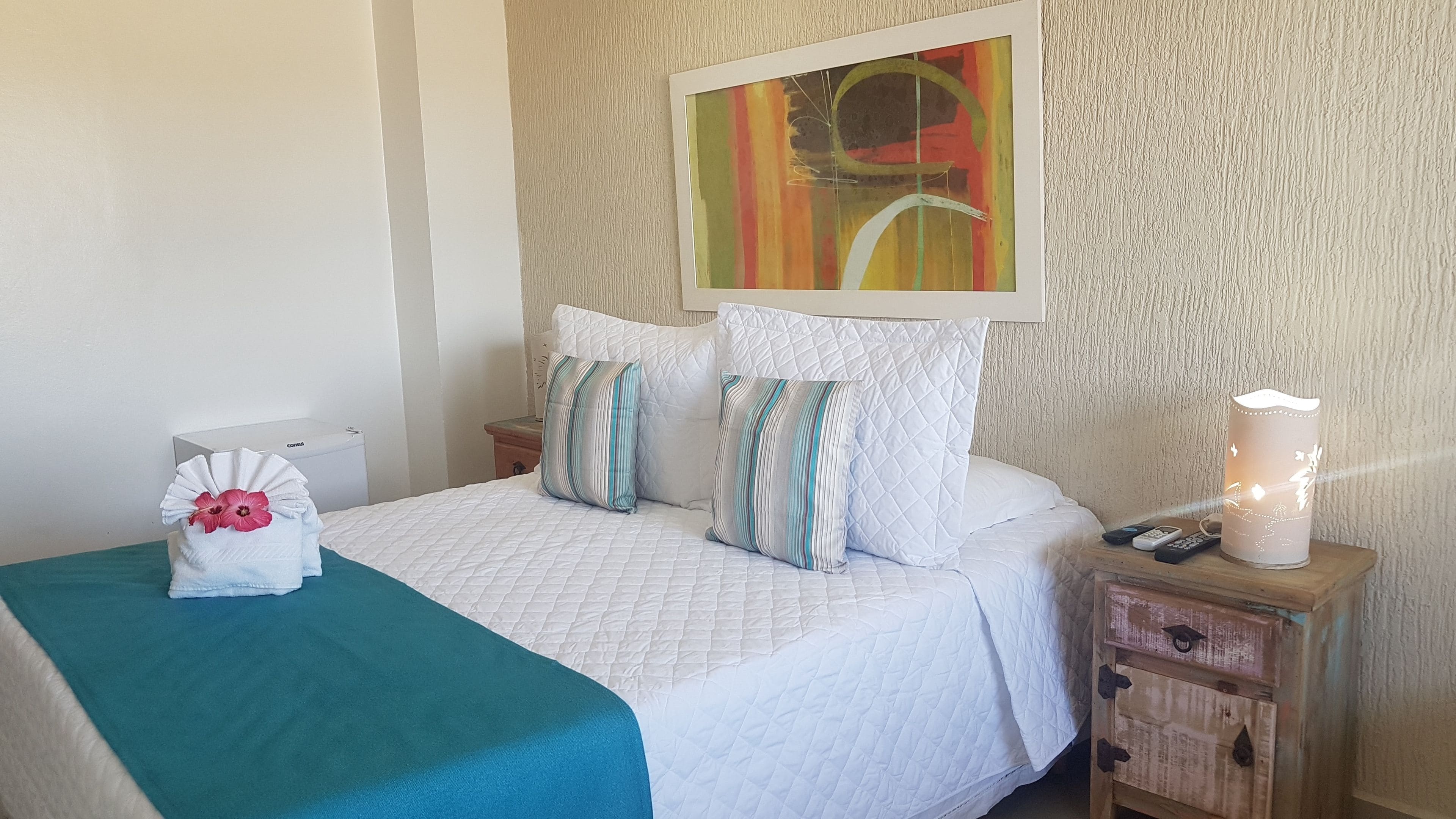 A neatly made bed with a teal blanket, white pillows, and a white quilted duvet. There is a painting above the bed, a bedside table with a lamp, and a small wooden cabinet.