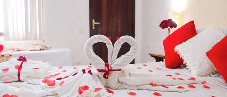 Una cama cuidadosamente arreglada con pétalos de rosa esparcidos sobre las sábanas, una toalla dispuesta en forma de corazón y una almohada roja.