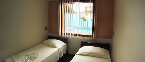 A small hotel room with two single beds, a window with blinds, and a view of a pool.