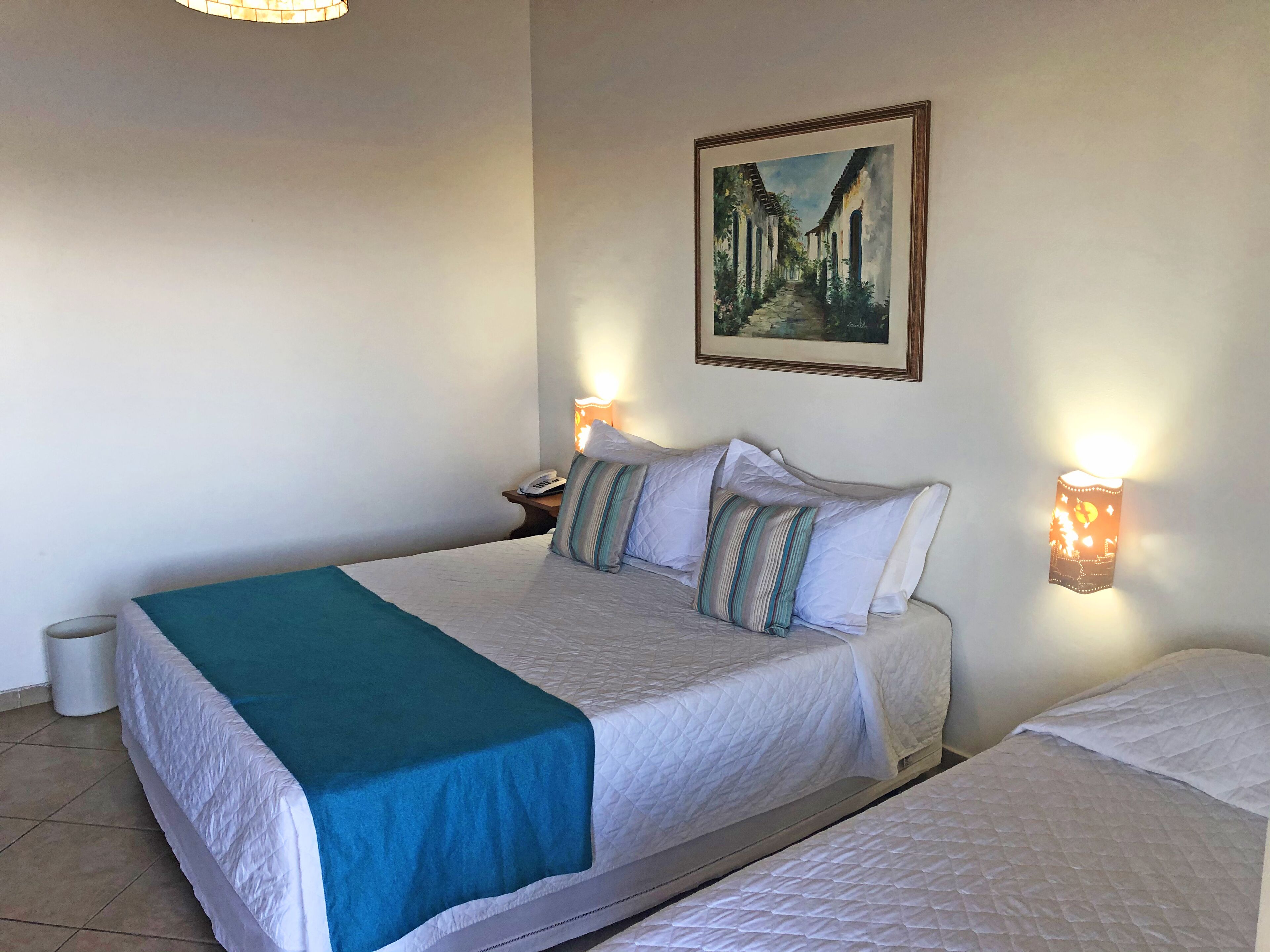 A hotel room with a bed, bedside table, lamp, and a painting on the wall.