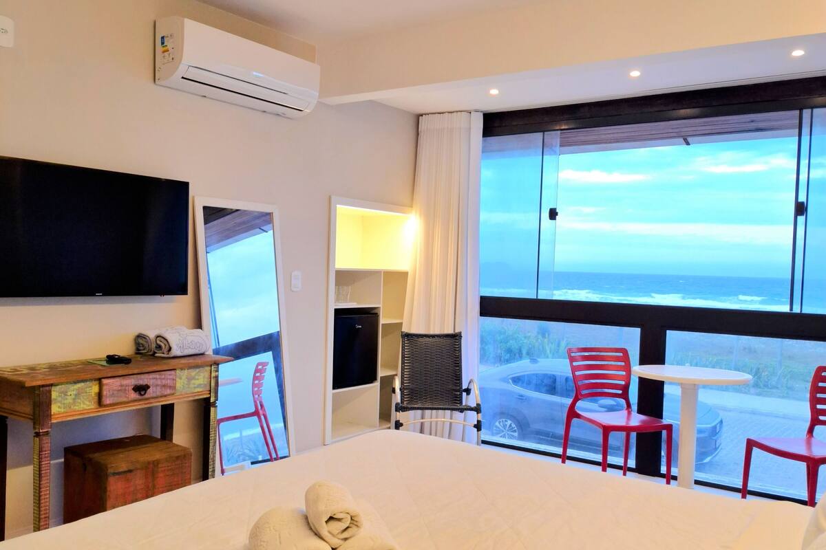 A hotel room with a bed, a desk, a TV, a window with a view of the ocean, and red chairs.