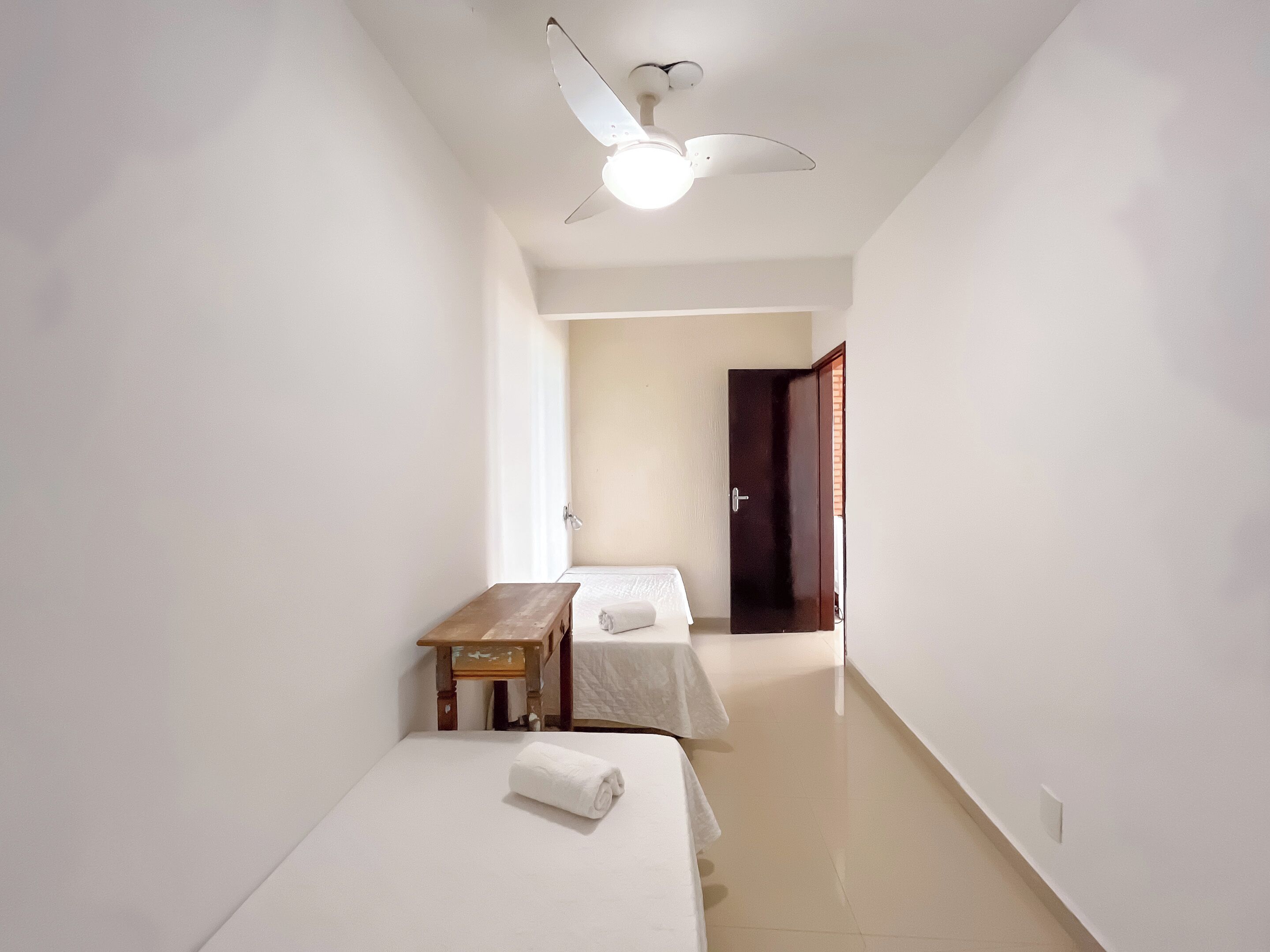 A narrow, well-lit room with two beds, a wooden bench, and a ceiling fan.