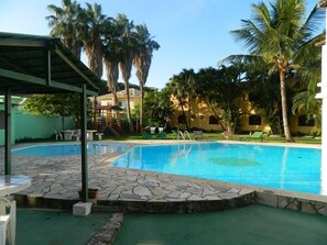Piscina com ladrilhos de pedra, rodeada por palmeiras e um espaço coberto com assentos.