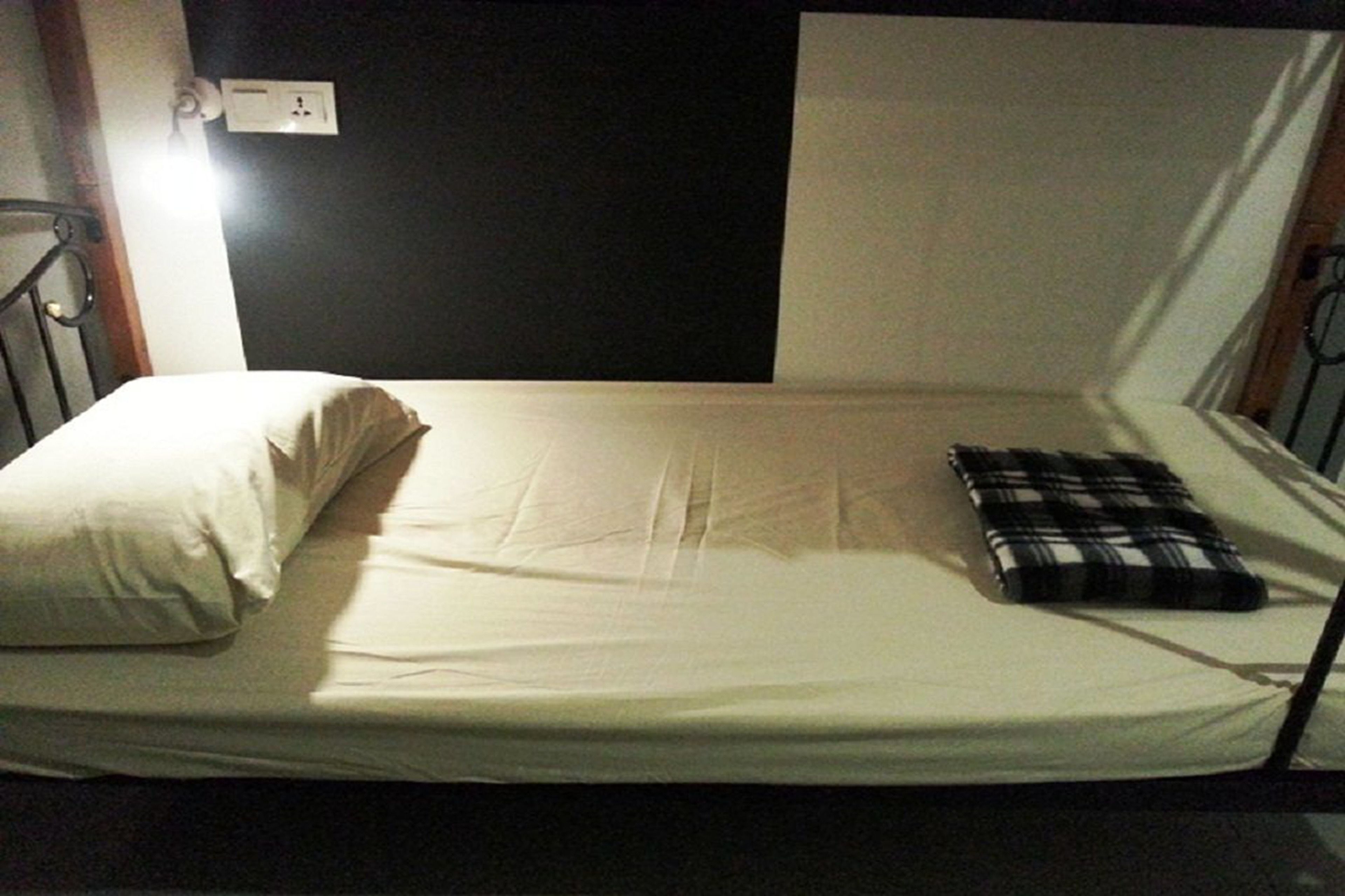A single bed with a white mattress, a black and white checkered pillow, and a plain wall with a power outlet.
