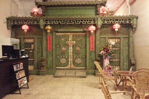 Een traditionele Chinese poort met ingewikkelde decoraties en rode versieringen, een receptiebalie met tijdschriften, en rieten stoelen op de voorgrond.