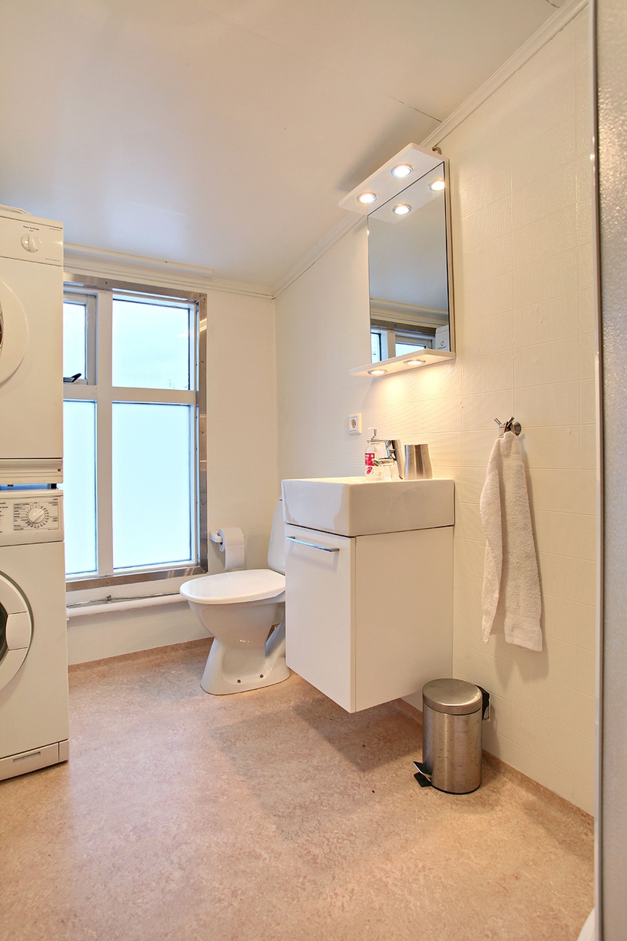 A small, clean bathroom with a toilet, sink, and mirror. There is a window providing natural light and a trash can next to the sink.