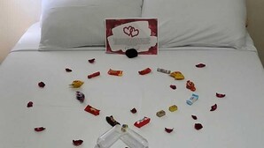 Una cama con sábanas blancas, una tarjeta roja y blanca, y pétalos de rosa y bombones esparcidos.