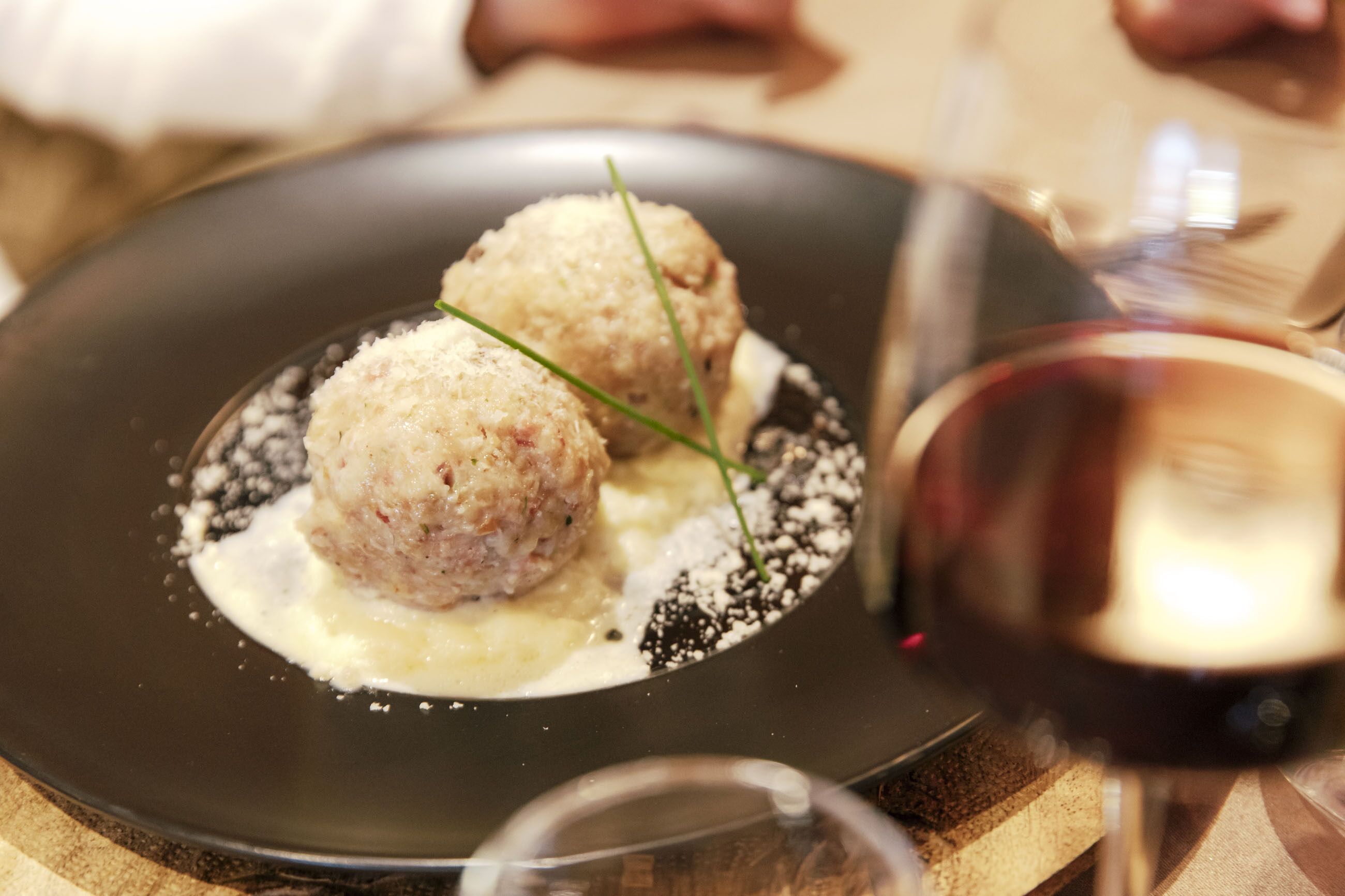 Une assiette contenant deux portions de nourriture de forme ronde, peut-être des boulettes de viande, sur une assiette foncée, le tout nappé d’une sauce blanche et agrémenté d’une garniture.