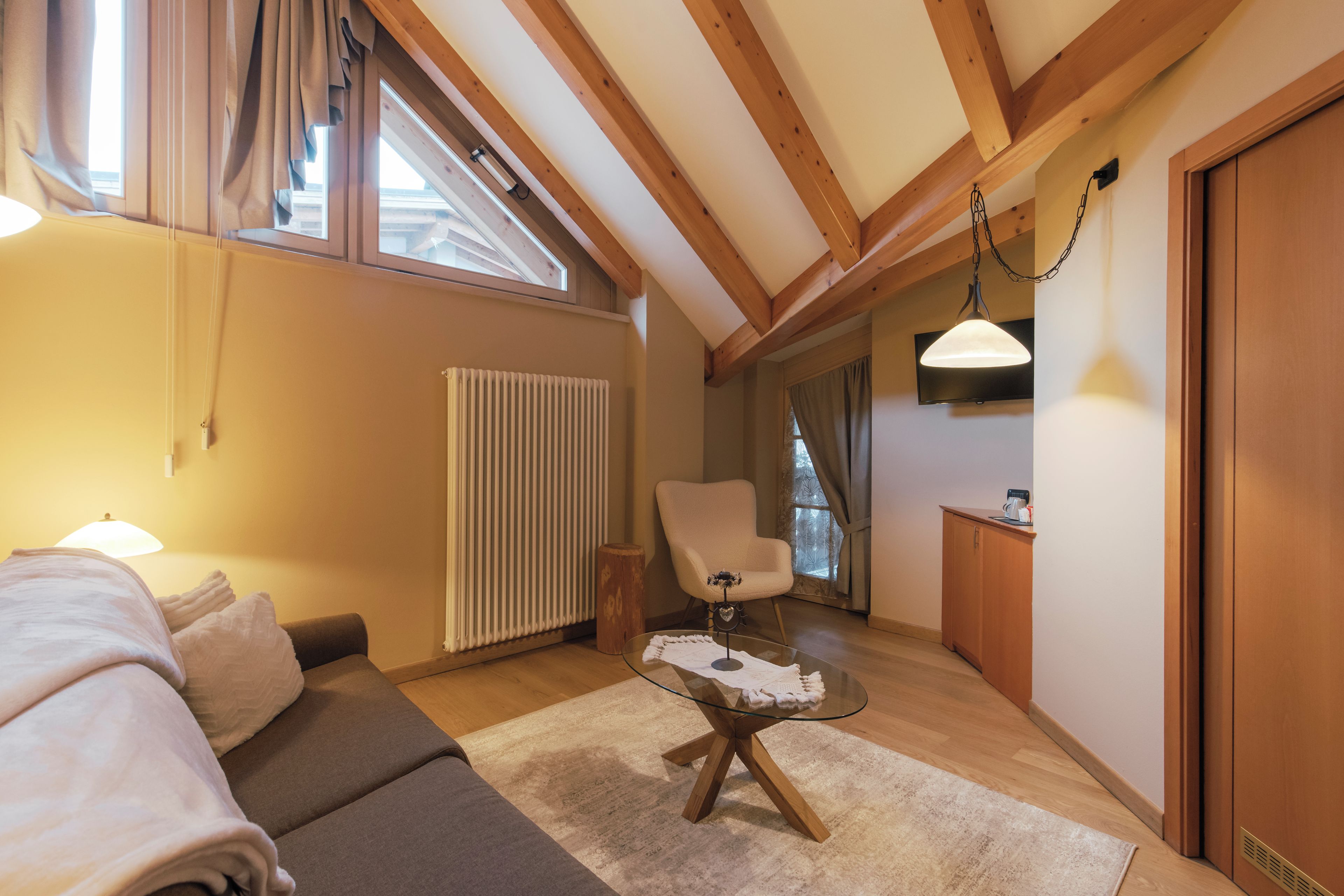 A cozy room with a sofa, a glass coffee table, a chair, a radiator, and a wooden ceiling.