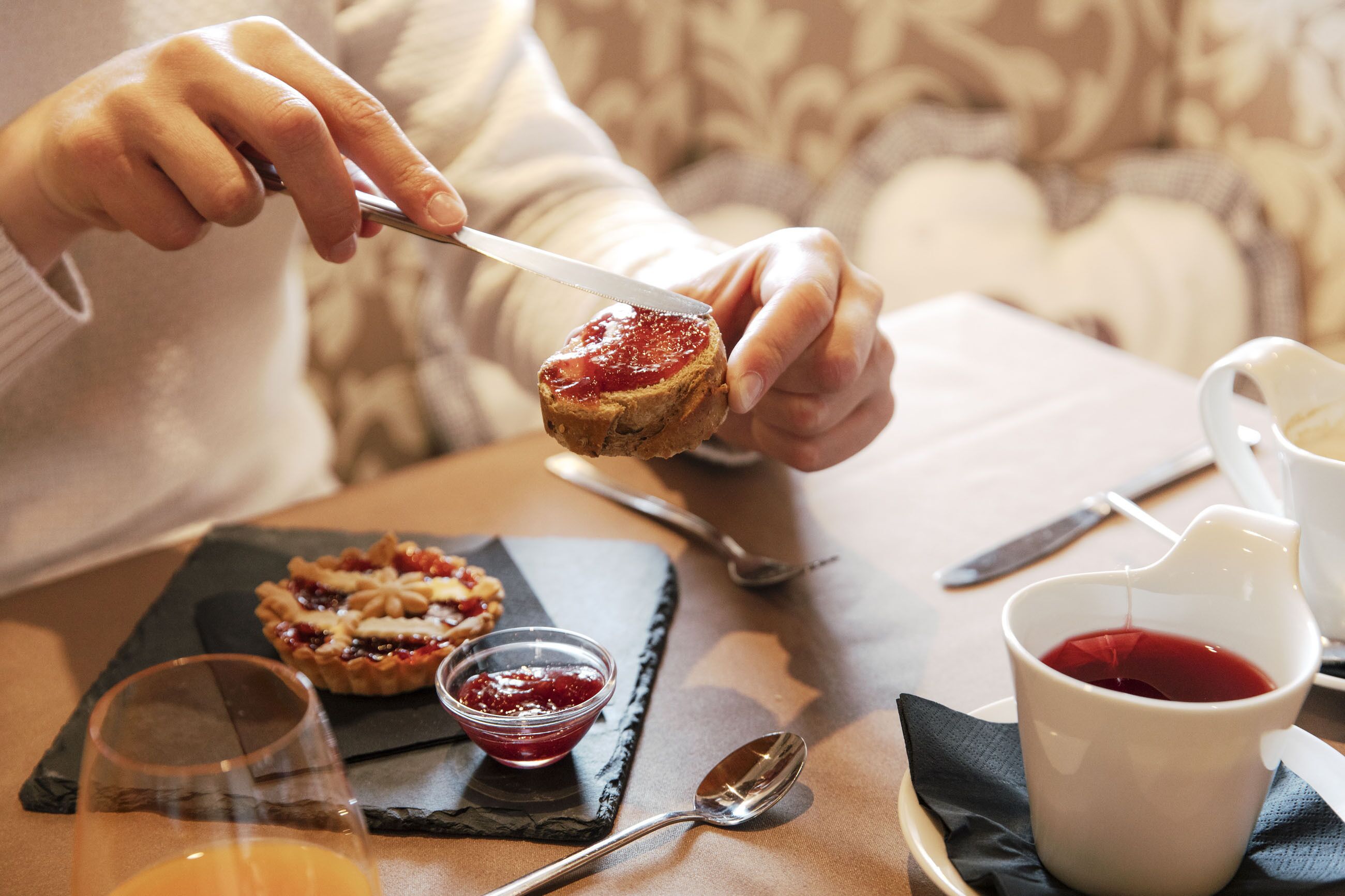 Una persona untando mermelada en una tostada, un cuenco pequeño de mermelada, un vaso de jugo de naranja y una taza blanca con líquido rojo.