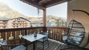Um deck de madeira com mesa e cadeiras, uma cadeira suspensa e vista para montanhas e edifícios.