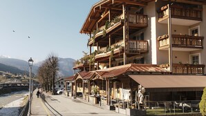 A multi-story building with balconies, a covered outdoor seating area, and a view of a river and mountains in the background. - Hotel Astoria  (Primiero San Martino di Castrozza)