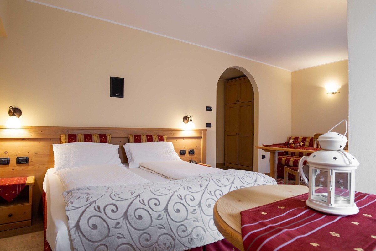Una habitación de hotel ordenada con una cama, mesitas de noche, una pequeña mesa con una silla y un farol decorativo.