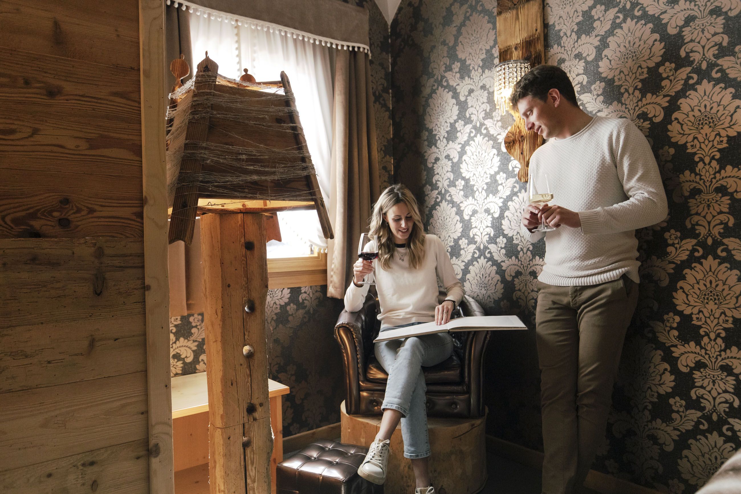 A room with a rustic wooden interior, a person sitting on a leather chair reading a book, and another person standing holding a glass.