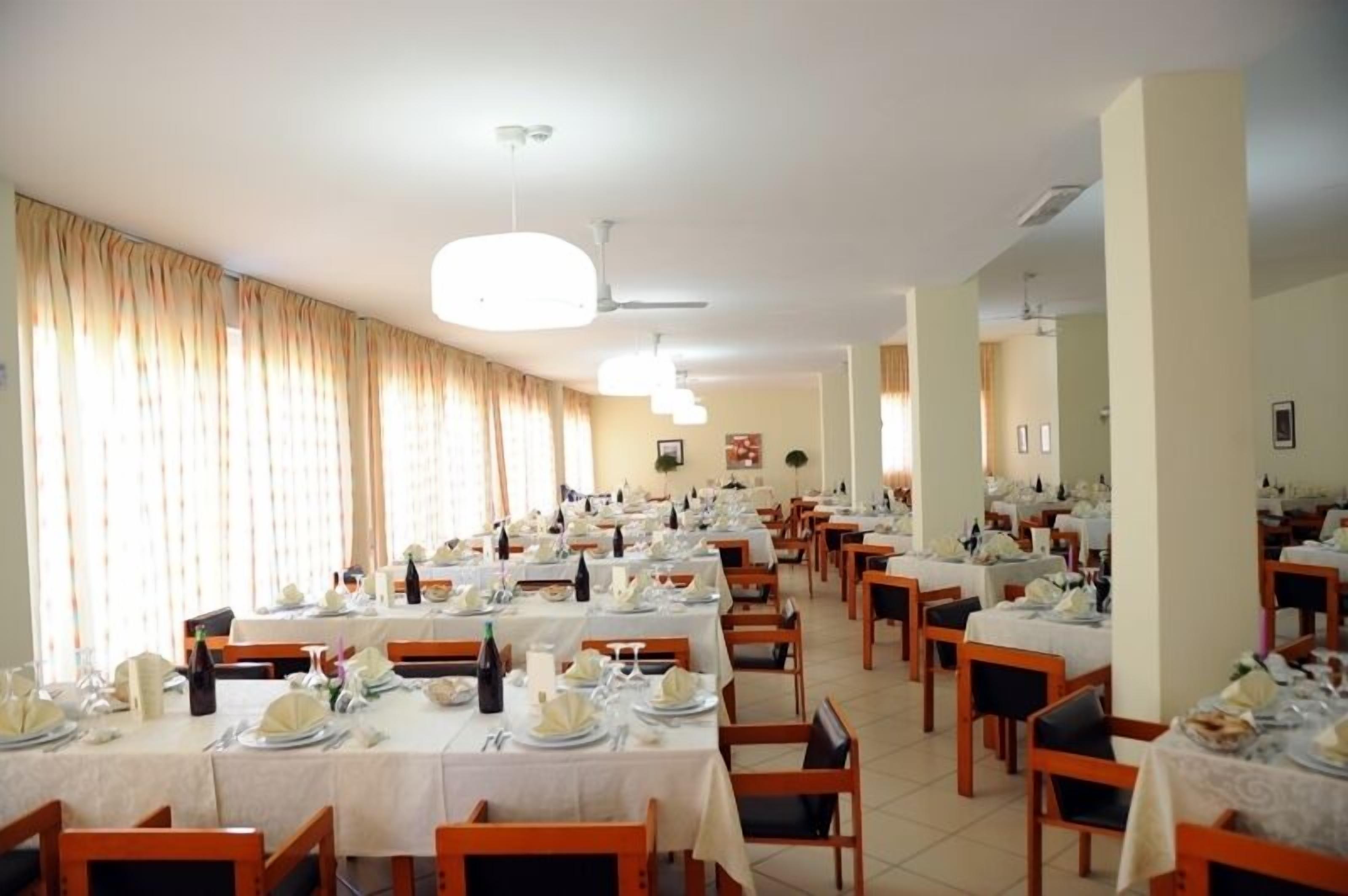 Un'ampia sala da pranzo con numerosi tavoli apparecchiati, ventilatori a soffitto e tende alle finestre.