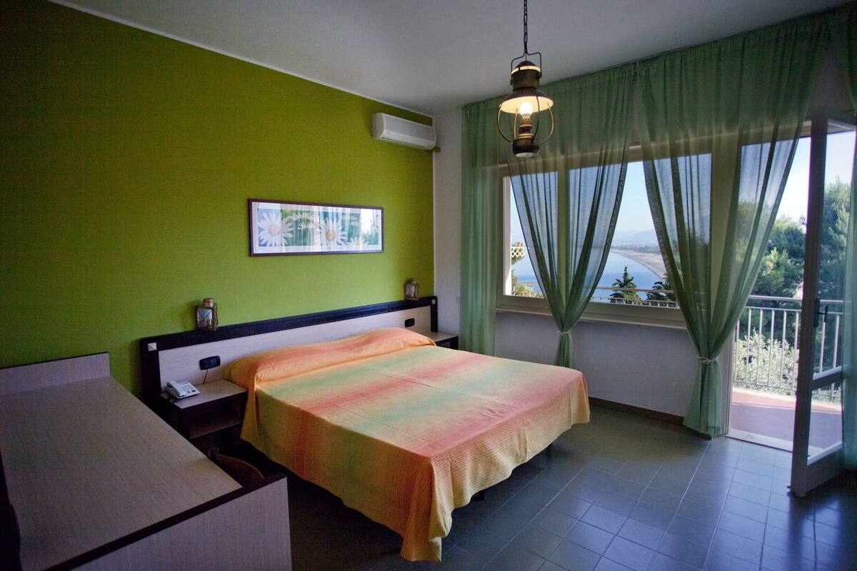 A hotel room with a bed, a desk, a chair, a window with curtains, and a view of the sea.