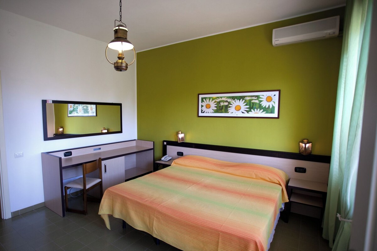 Habitación de hotel con cama, escritorio, silla, espejo, mesita de noche y aire acondicionado instalado en la pared.