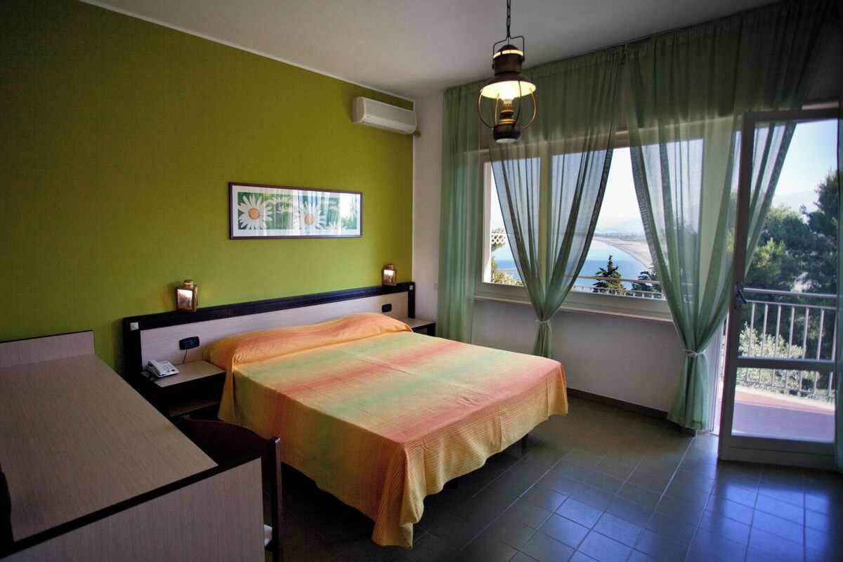 A bedroom with a bed, a nightstand, a lamp, a picture on the wall, and a window with a view.