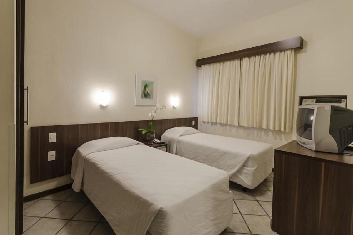 Una habitación de hotel con dos camas, un escritorio con televisor y una ventana con cortinas.