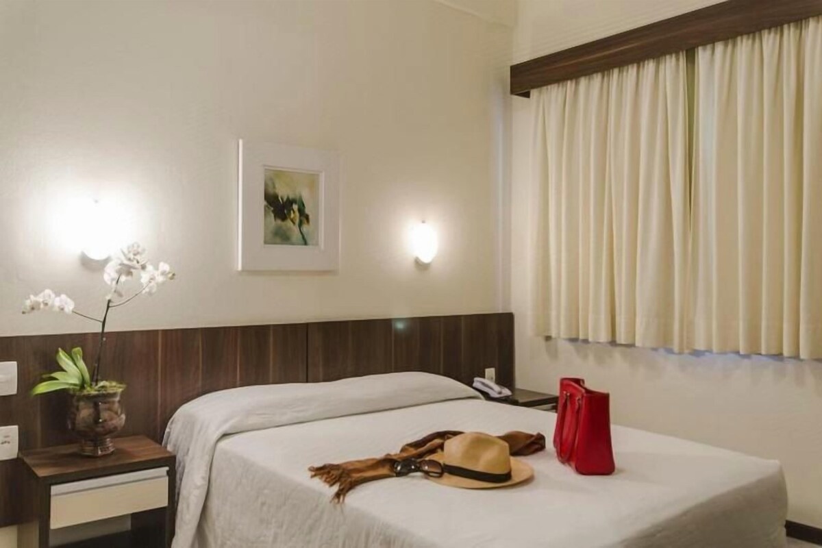 Una habitación de hotel con una cama, una mesita de noche, un florero con flores, un cuadro en la pared y una ventana con cortinas.