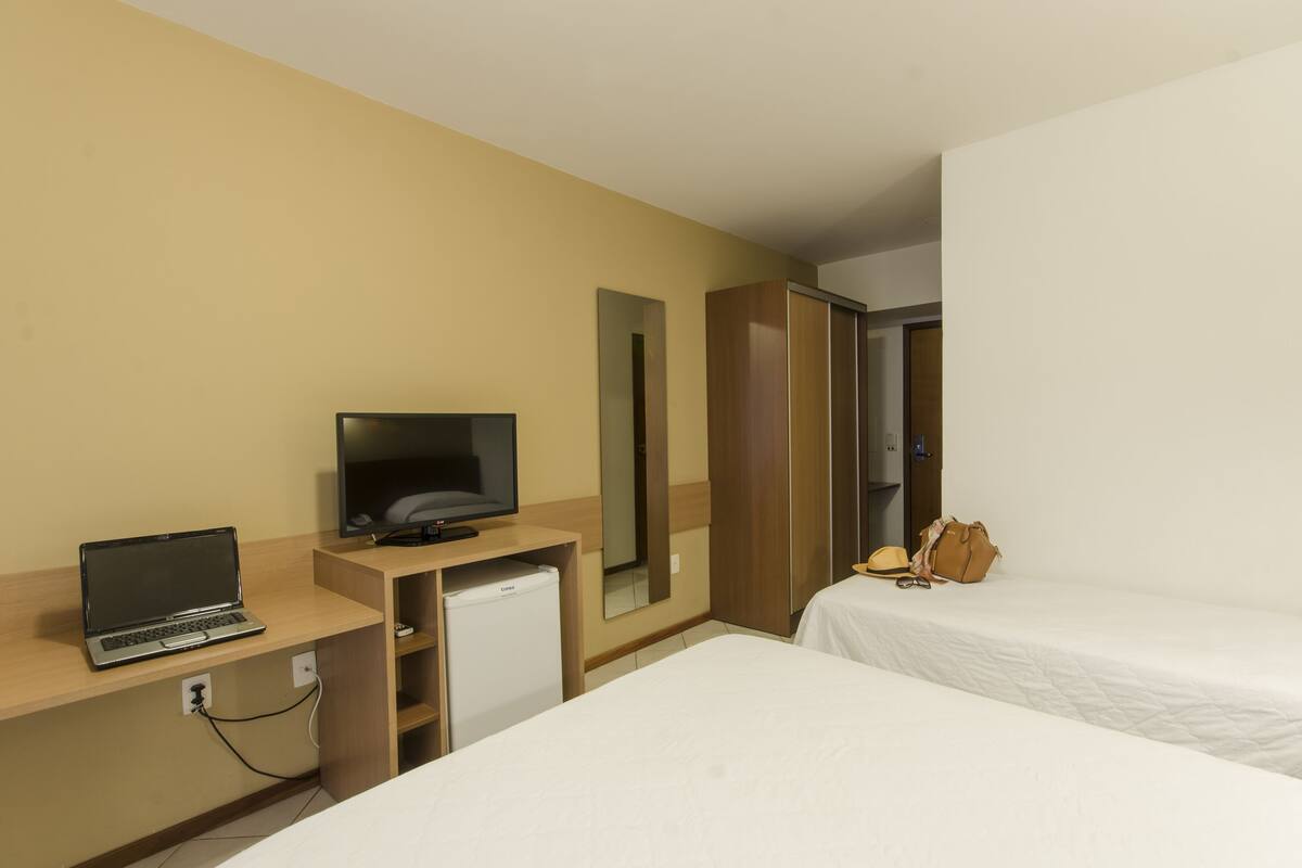 A hotel room with a bed, a desk with a laptop, a television, a mirror, and a wardrobe.