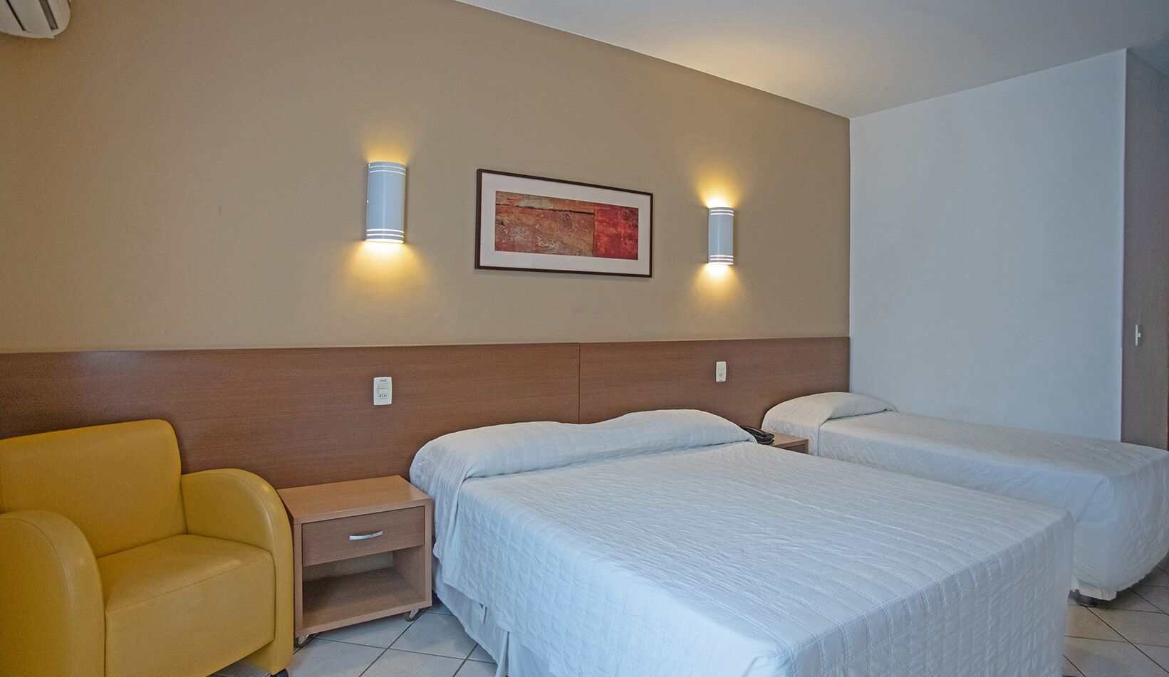 A hotel room with two beds, a nightstand, a chair, and wall-mounted lights.