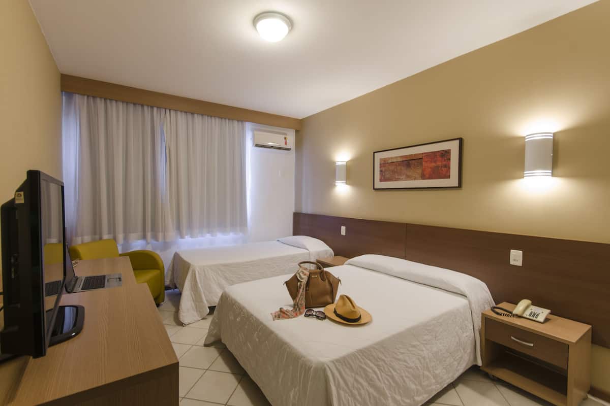 A hotel room with two beds, a desk with a computer, a chair, a bedside table with a phone, and a wall-mounted light.