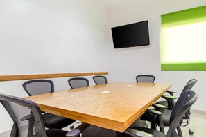 Un salón de conferencias con una mesa de madera, seis sillas, un televisor de pantalla plana en la pared y un ventanal con persiana verde.