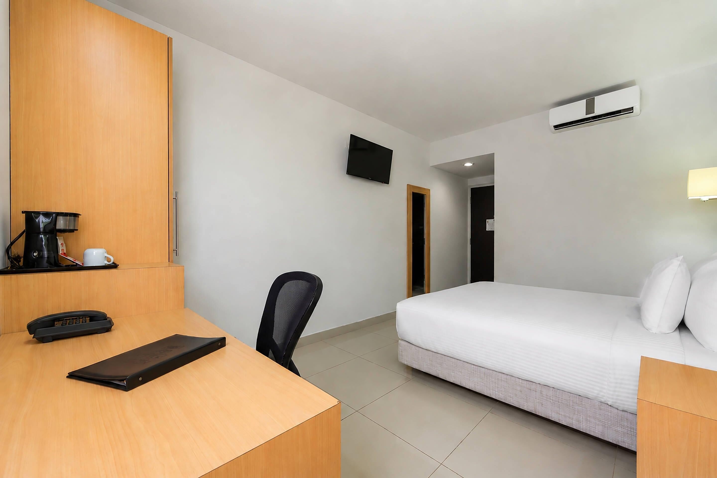 Habitación de hotel con escritorio de madera, una silla, cama con ropa blanca, televisor montado en la pared y unidad de aire acondicionado.