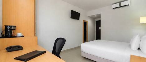 Habitación de hotel con escritorio de madera, una silla, cama con ropa blanca, televisor montado en la pared y unidad de aire acondicionado.