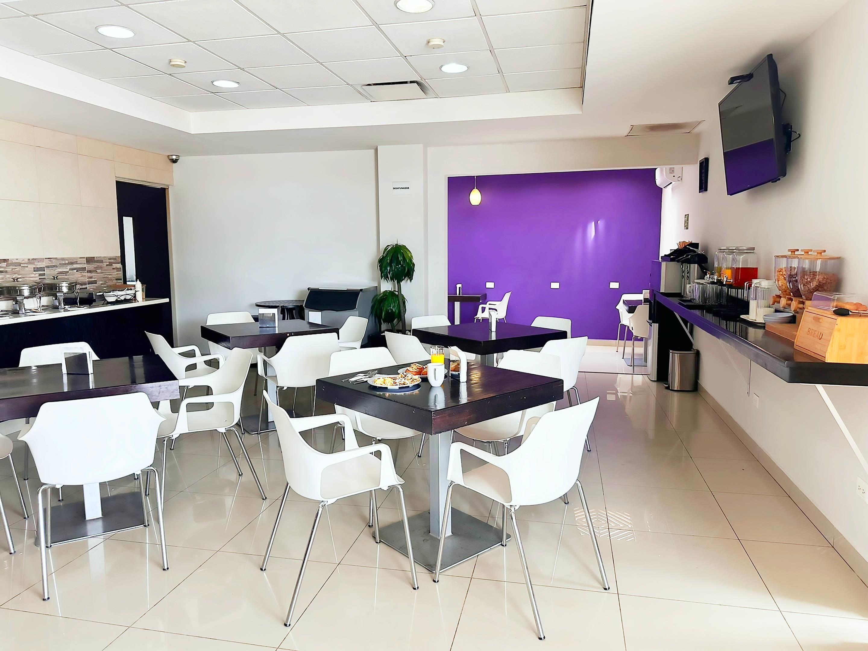 Un comedor moderno con sillas y mesas blancas, una pared de acento morada y un mostrador de buffet con comida y bebidas.