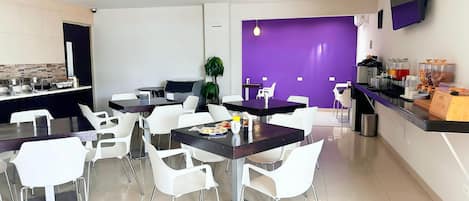 Un comedor moderno con sillas y mesas blancas, una pared de acento morada y un mostrador de buffet con comida y bebidas.