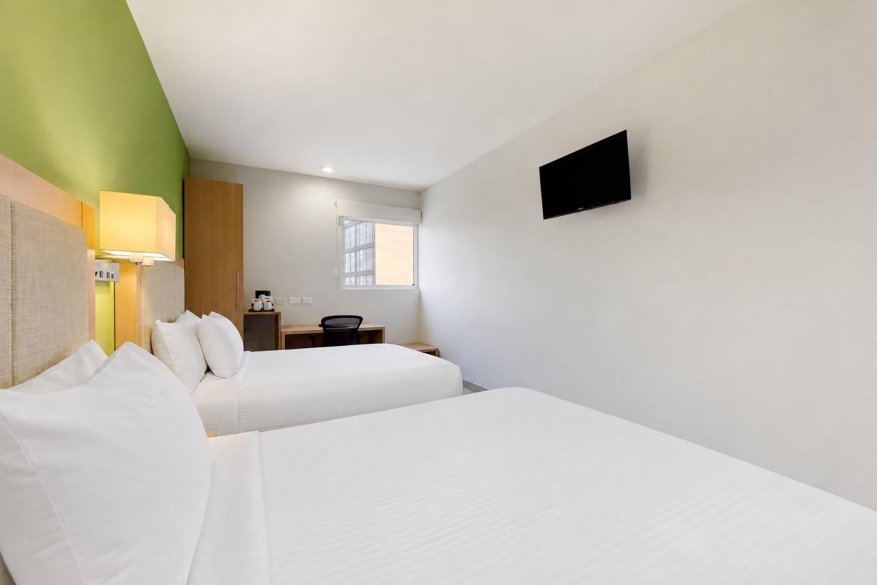 A hotel room with two beds, a desk, a chair, a TV mounted on the wall, and a window.