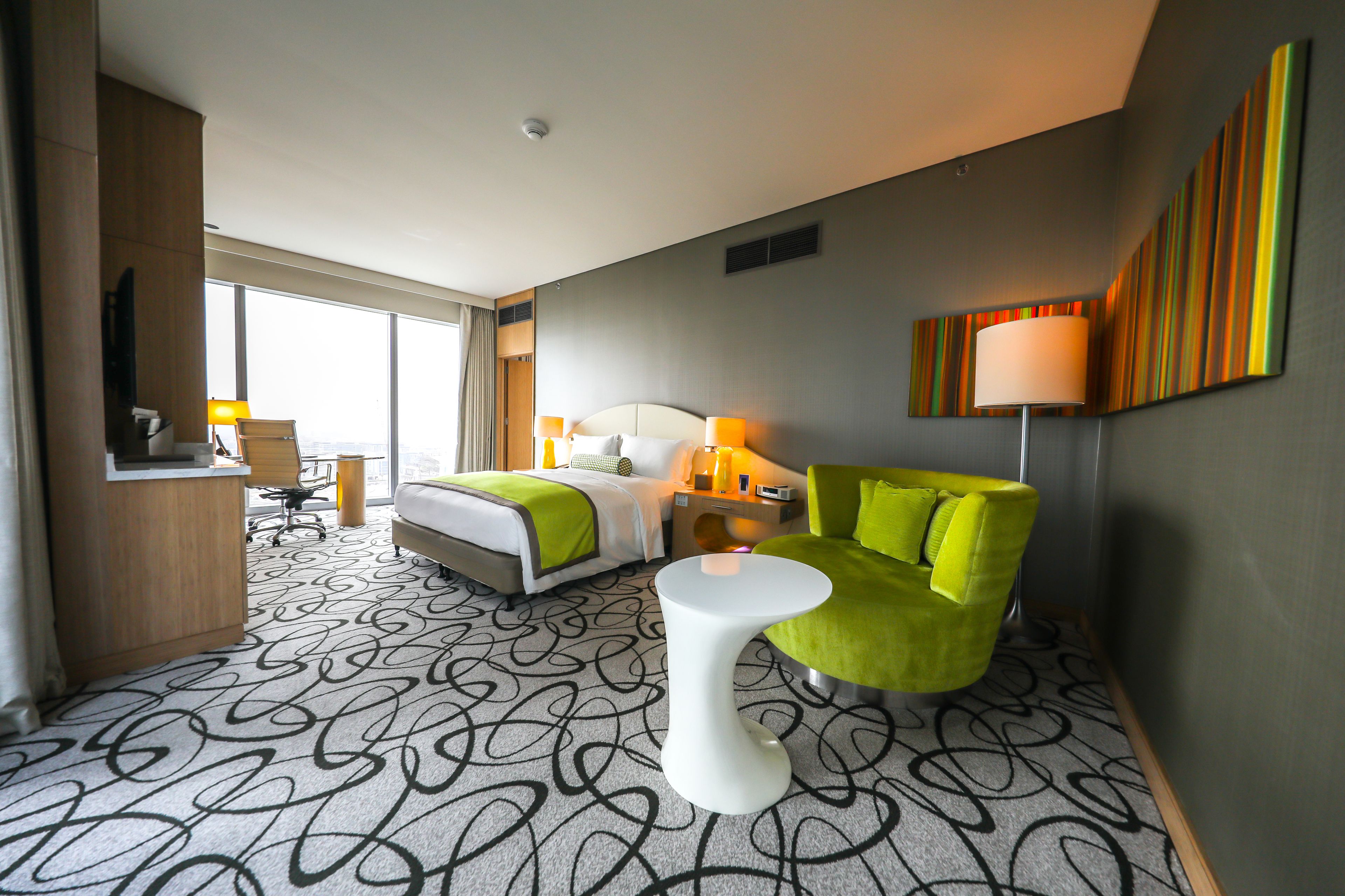 A hotel room with a bed, a green armchair, a white side table, and a desk with a chair.
