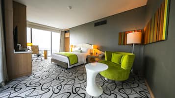 A hotel room with a bed, a green armchair, a white side table, and a desk with a chair.