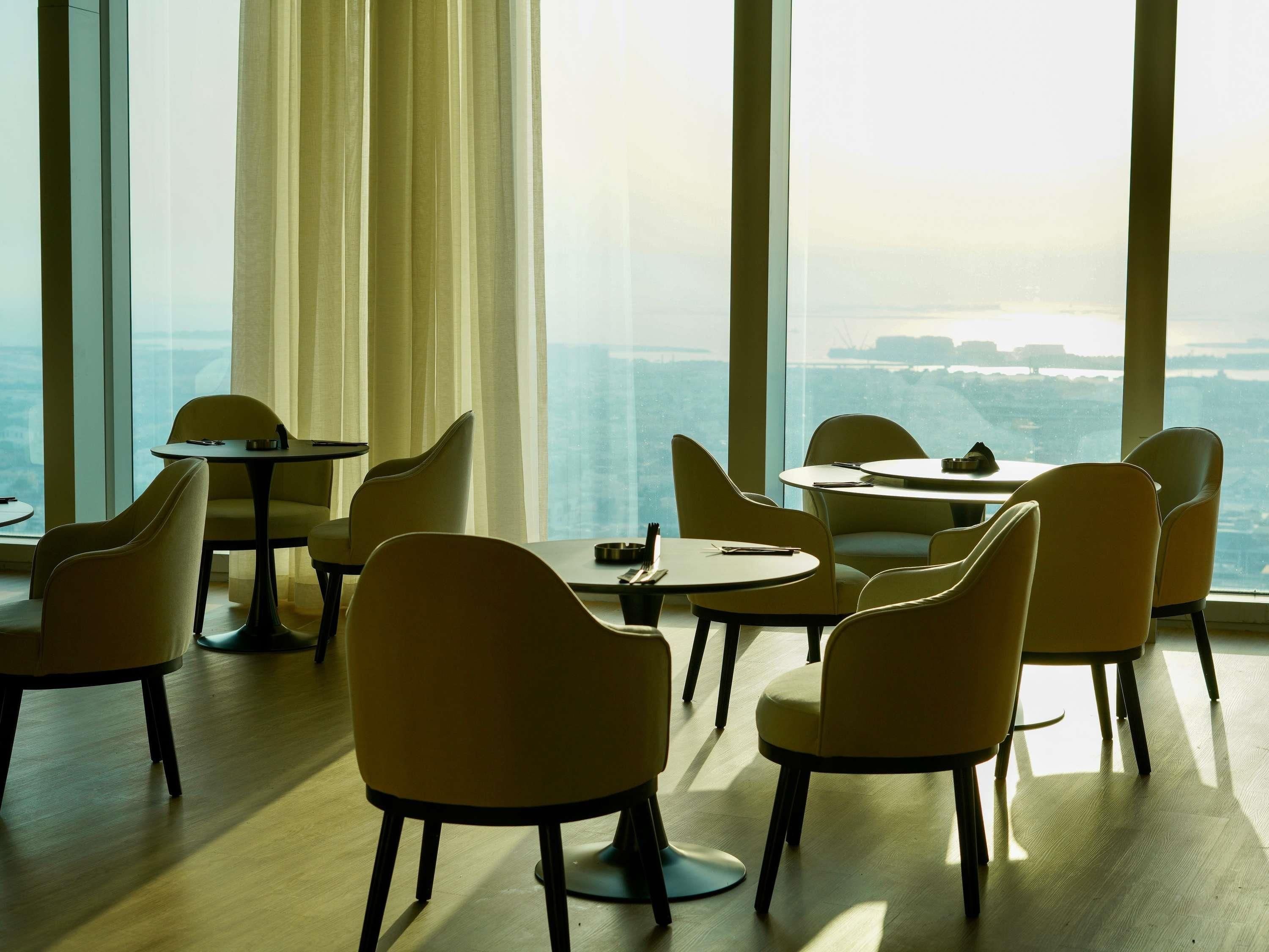A modern dining area with round tables and chairs, large windows with curtains, and a view of a cityscape.