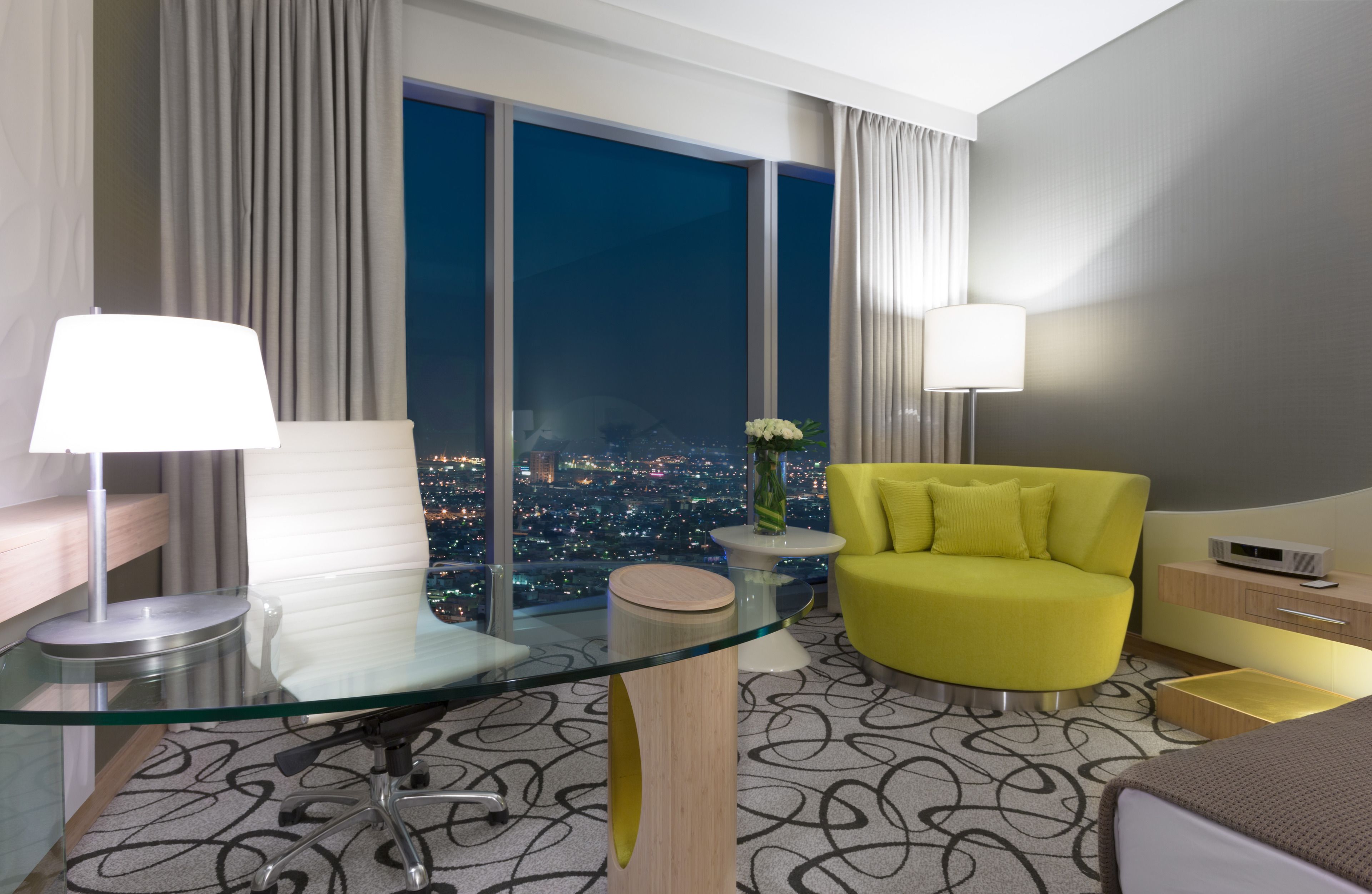 A modern hotel room with a glass desk, a yellow armchair, a city view, and a large window.