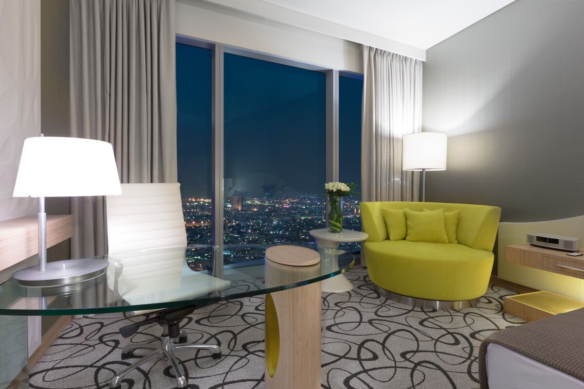 A modern hotel room with a glass desk, a yellow armchair, a city view, and a large window.