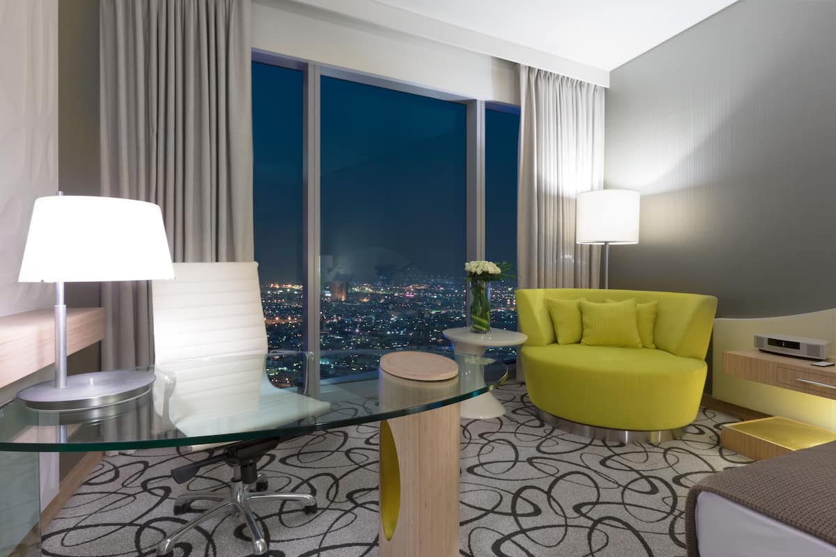 A modern hotel room with a glass desk, a yellow armchair, a city view, and a large window.