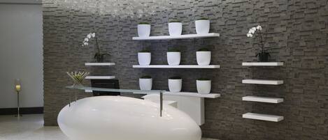 A modern interior with a central white reception desk, multiple white shelves with potted plants, and a hanging light fixture.